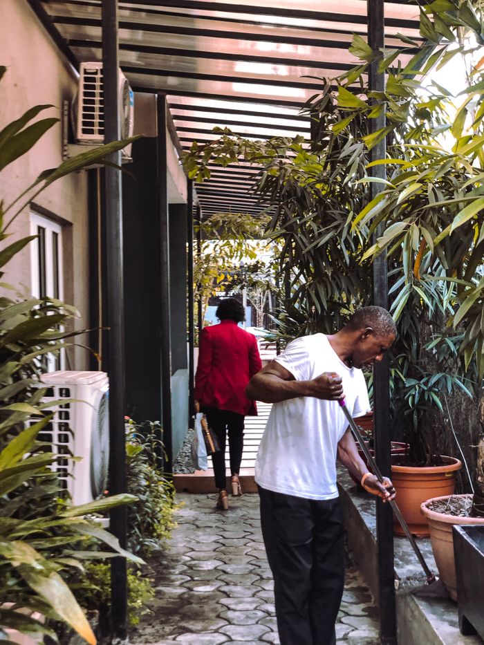 why-choose-us-03 A man sweeps an urban courtyard filled with lush tropical plants while a woman walks by.